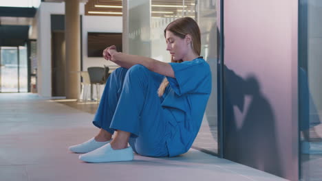 tired and overworked medical worker in scrubs sitting on floor of hospital corridor during break