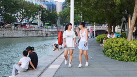 Two young women walk and chat beside a riverside promenade, passing seated people in a lively urban park with greenery and water, under soft daylight