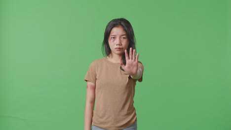 young asian woman with bruise on face and arms looking into camera showing hand sign to stop violence on green screen background in the studio