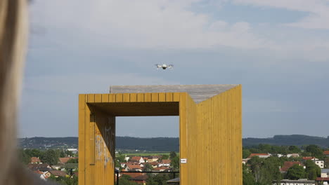 quadcopter consumer drone flying above structure in city suburbs in front of young woman, back view, slow motion