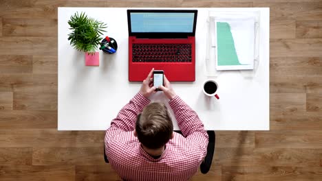 young man texting on phone, topshot, sitting behind desk with laptop and coffee