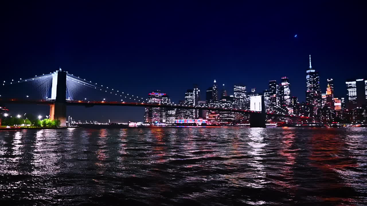 Dark waterscape of the river reflects multiple city lights of metropolis. Low angle view at the Brooklyn Bridge and New York skyline at night