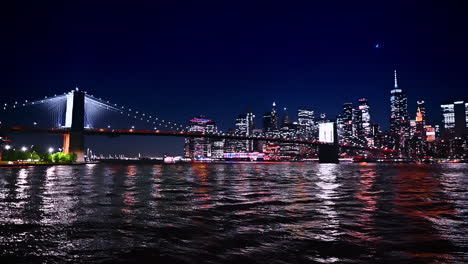 dark waterscape of the river reflects multiple city lights of metropolis. low angle view at the brooklyn bridge and new york skyline at night