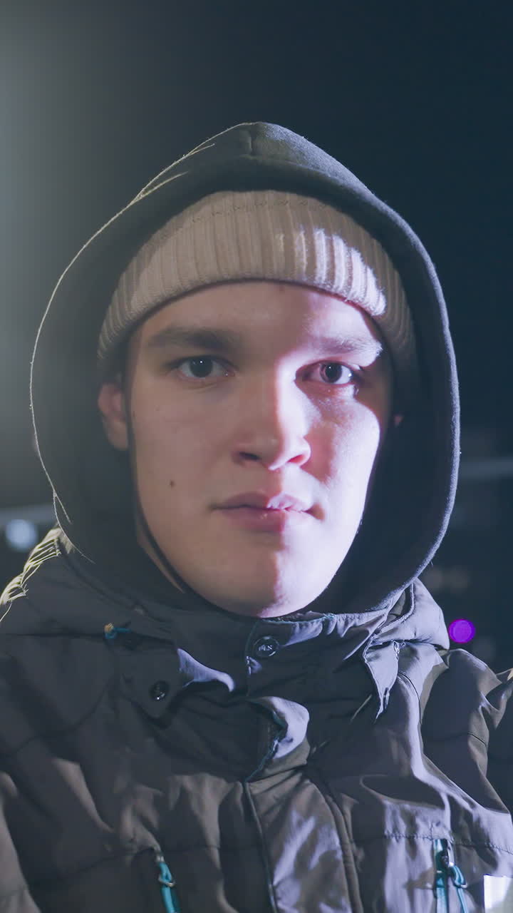 niño joven con capucha sosteniendo la pelota en la mano, mirando pensativo durante la práctica nocturna bajo la luz de la calle brillante con el paisaje urbano en el fondo