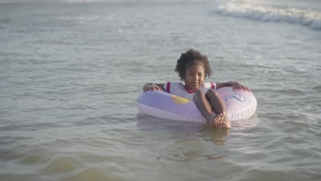 Happy-Afro-American-young-girl-smiling-at-beach,-floating-in-sea-with-float