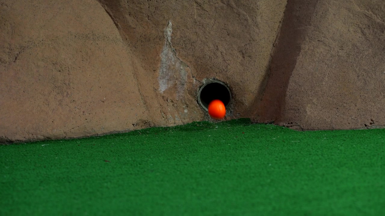 Una pallina da minigolf arancione esce da un tubo in un muro e rotola mancando la buca