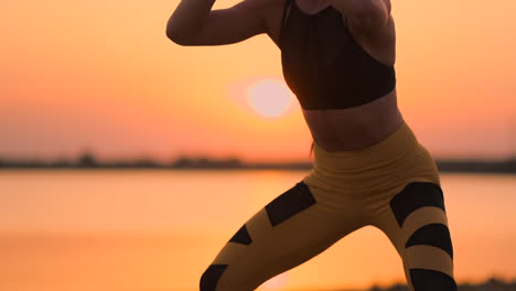 Vista-Lateral-De-Una-Mujer-Joven-Haciendo-Sentadillas-Al-Aire-Libre.-Vista-Lateral-De-Una-Joven-Mujer-Fitness-Haciendo-Sentadillas-De-Pie-Junto-Al-Lago-En-La-Arena-Al-Atardecer-En-Cámara-Lenta