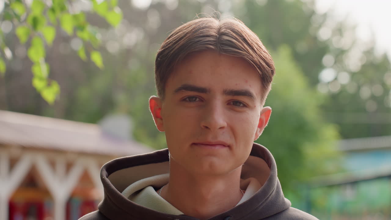 Blanke tienerjongen met hoodie buiten, peinzend portret bij houten tuinprieel met berkenbladeren en zacht zonlicht. Student kijkt bedachtzaam, aspirant-muzikant bekijkt de omgeving, casual model oefent