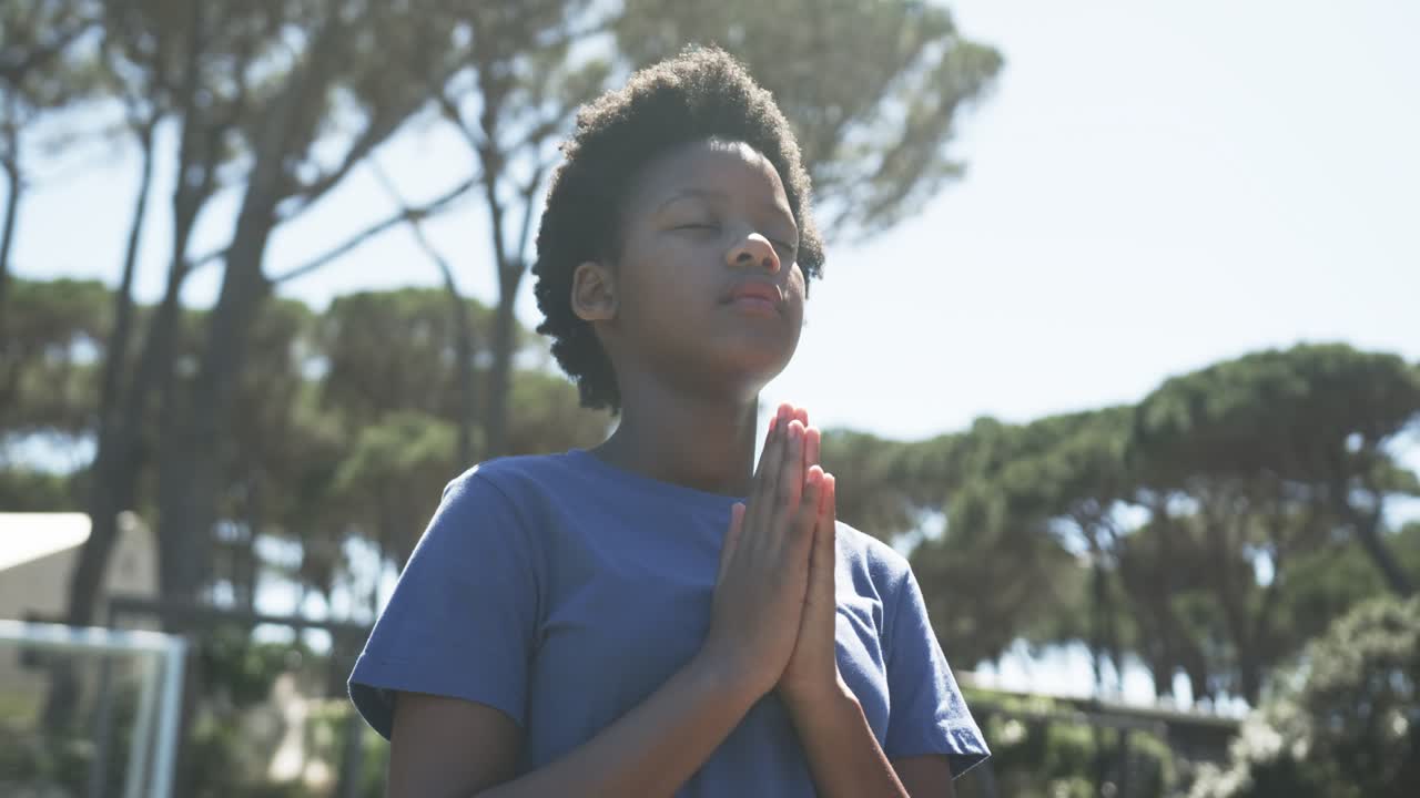 Giovane afroamericana in piedi in un parco con una maglietta blu, palmi delle mani uniti e occhi chiusi verso il sole