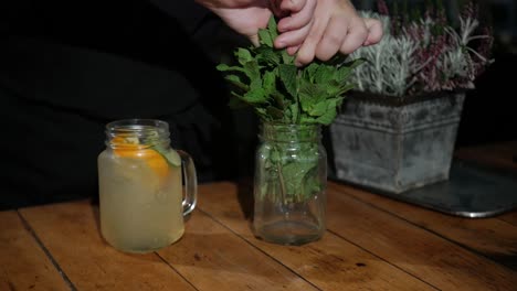 hand of barman adding fresh green mint into alcoholic cocktail on wooden table