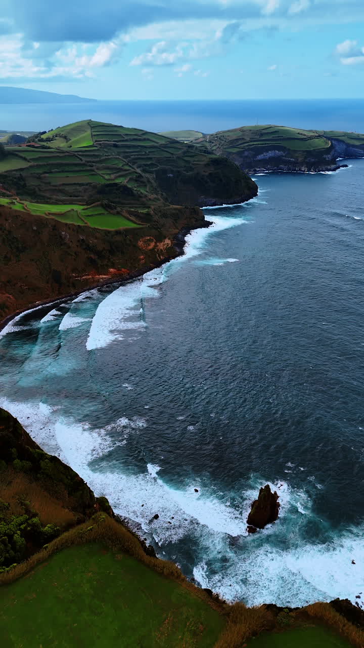 White foamy waves arrive to the rocky shore. Aerial perspective on the unusual mountains at the coast of the Azorean Islands. Vertical video.
