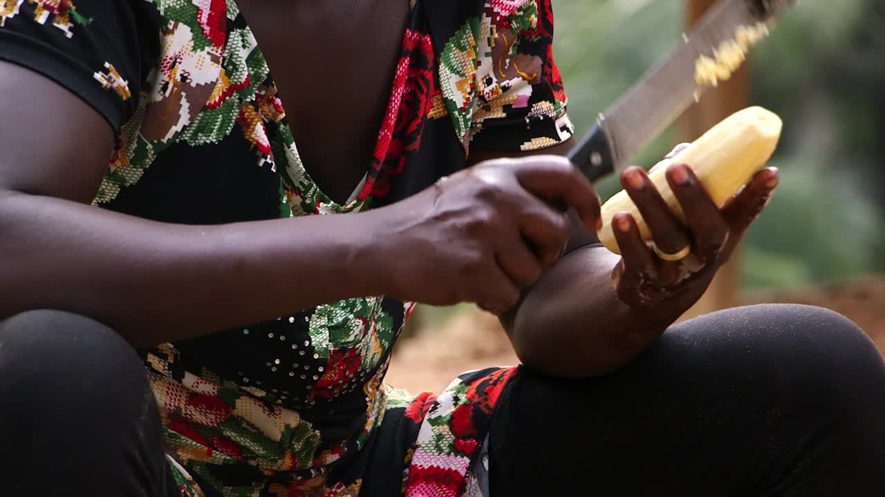 Primer plano de mujer ghanesa pelando yuca con un machete, preparación tradicional de alimentos al aire libre en la zona rural de África Occidental.