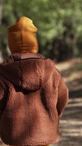 following the lovely baby boy wearing brown jacket and yellow cap. sweet kid on the walk in the autumn park. vertical video