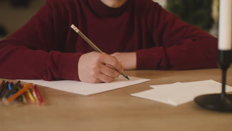 Vista-De-Cerca-De-Las-Manos-De-Un-Niño-Escribiendo-Una-Carta-De-Deseos-Sentado-En-Una-Mesa