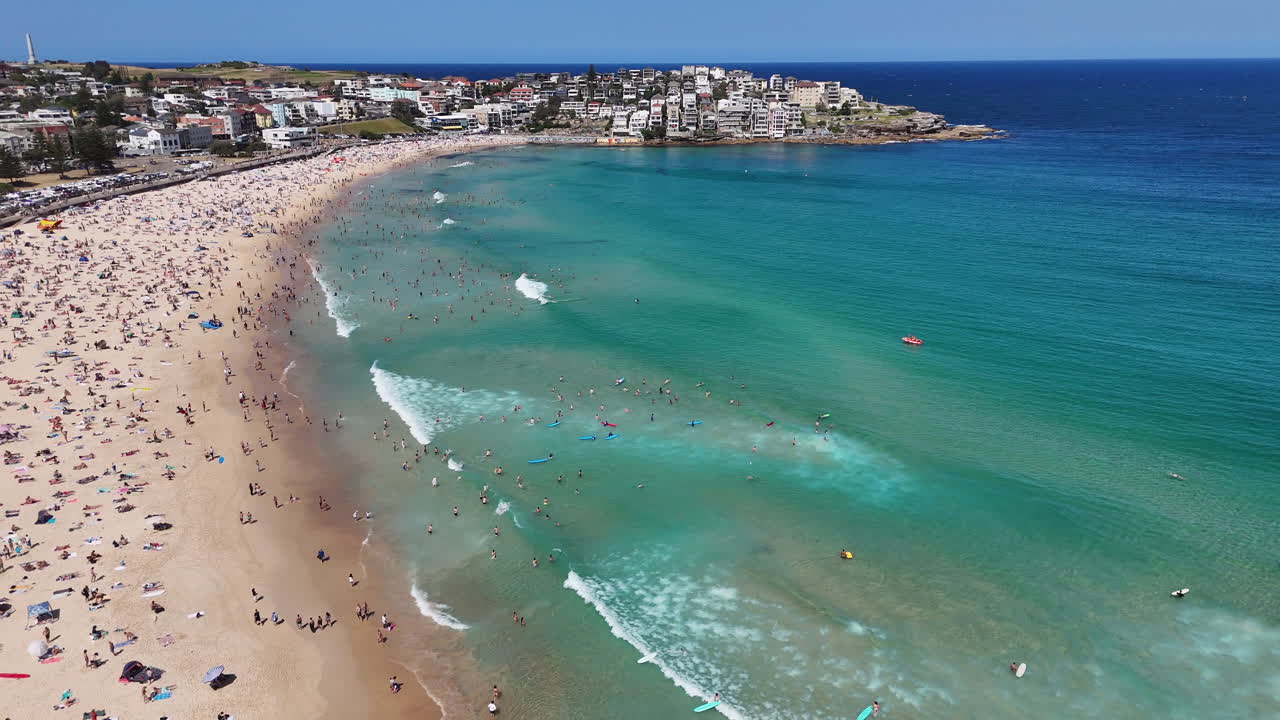 Panning drone shot of Bondi Beach, highlighting sunbathers, swimmers, and surfers along the famous Australian coast. Aerial footage.