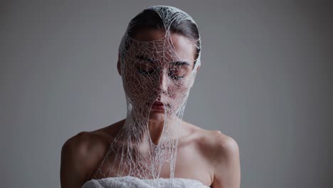 female model enveloped by white towel and spiderweb mesh. experiencing intense emotional struggle with closed eyes against minimalist gray studio background. conveying vulnerability and inner tension