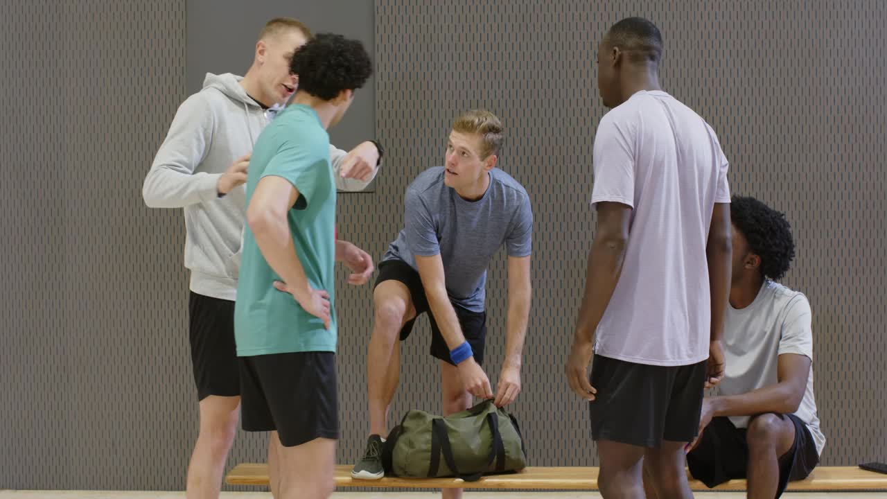 Männliches Team versammelt sich auf der Turnbank, nachdem die Sporttaschen abgestellt wurden; ein Mann in Blau-Grau öffnet seine Tasche und bindet die Schuhe für das Spiel.