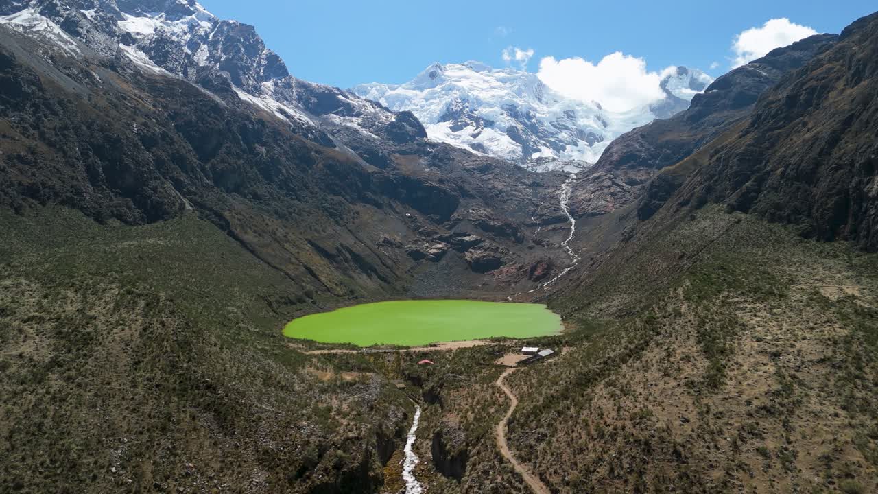 A stunning aerial drone shot flies forward, approaching the uniquely bright green waters of Laguna Shallap, a glacial lake nestled in a valley below snow-capped peaks in Peru's Andes