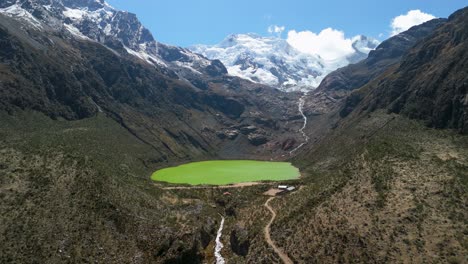 a stunning aerial drone shot flies forward, approaching the uniquely bright green waters of laguna shallap, a glacial lake nestled in a valley below snow-capped peaks in peru's andes