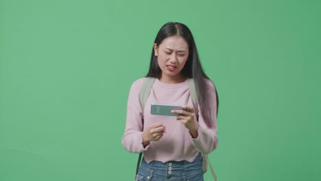 분노한 아시아 여성 학생이 모바일 게임을 하고 녹색 스크린 스튜디오에서 는 동안 잃습니다.