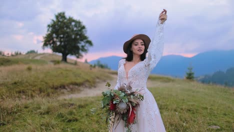 Gorgeous bride on mountains background. Beautiful young woman in hat and white dress with waving sleeves holding bouquet among nature.