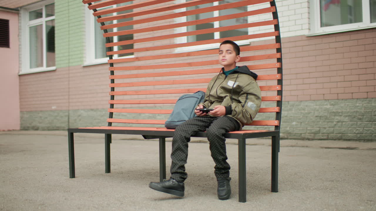 벽돌 벽과 창문이 있는 학교 야외 환경에서 한 어린 소년이 벤치에 앉아 가방을 옆에 두고 다리를 편안하게 움직이며 주변을 둘러보고 휴대폰 게임을 하고 있어요.
