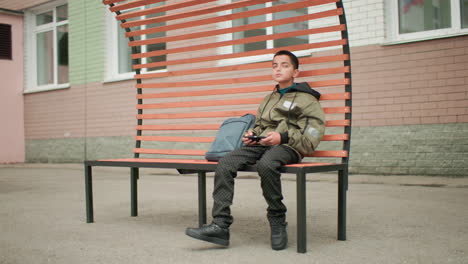 young boy seated on bench with bag by side moving leg casually while looking around and playing game on phone outdoor school environment with brick wall and windows background