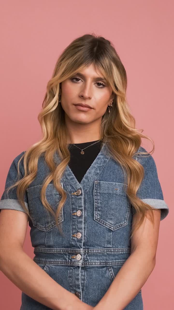 Confident transgender woman in denim dress against pink background