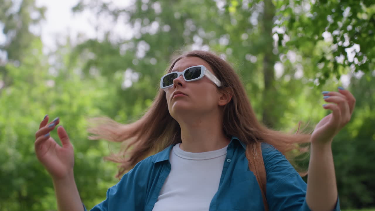 Student carefully puts on sunglasses in bright green park, adjusting frames with focused expression, wearing blue shirt and white top, surrounded by lush foliage under soft daylight