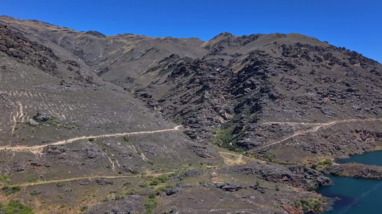On a bright sunny day, the drone flies towards the Dunstan Trail and Clutha River, approaching and revealing the bridge along the scenic trail in Central Otago, New Zealand