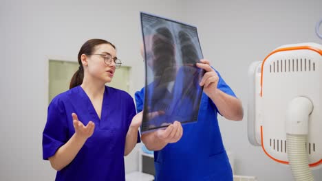 Un-Médico-Experimentado,-Un-Anciano-Con-Gafas-Y-Uniforme-Azul,-Se-Comunica-Y-Consulta-Con-Una-Joven-Morena,-Un-Médico-Con-Gafas,-Mientras-Realiza-Los-Resultados-De-La-Fluorografía-En-Una-Clínica-Moderna.