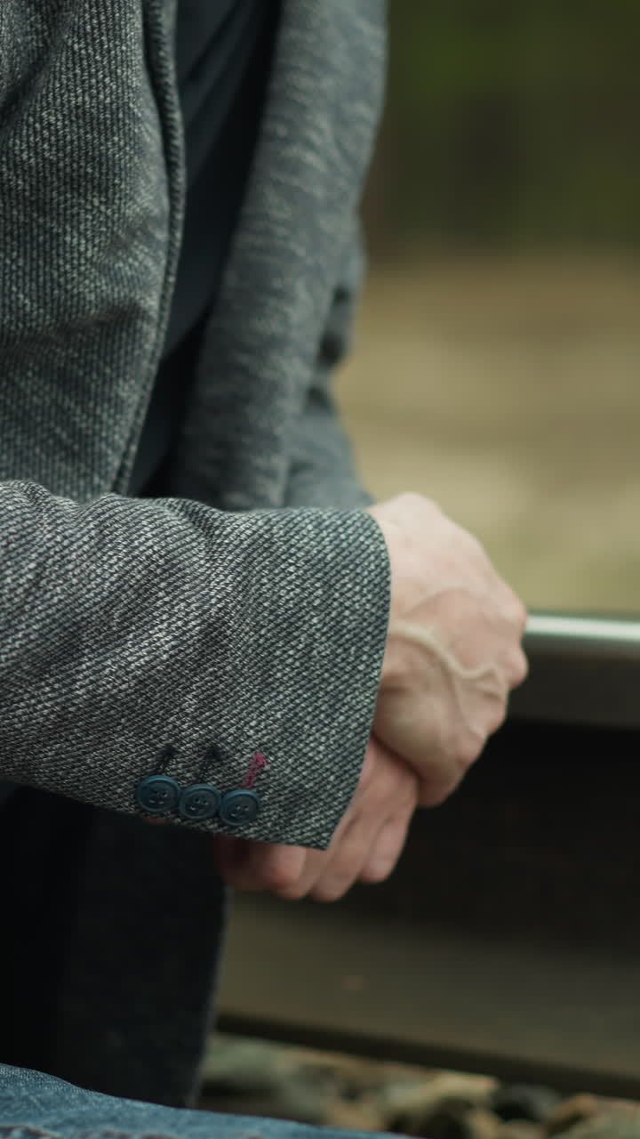 una vista de cerca de un hombre con un blazer gris y pantalones vaqueros azules, sentado y con las manos juntas en una vía ferroviaria, mientras la cámara se aleja de él