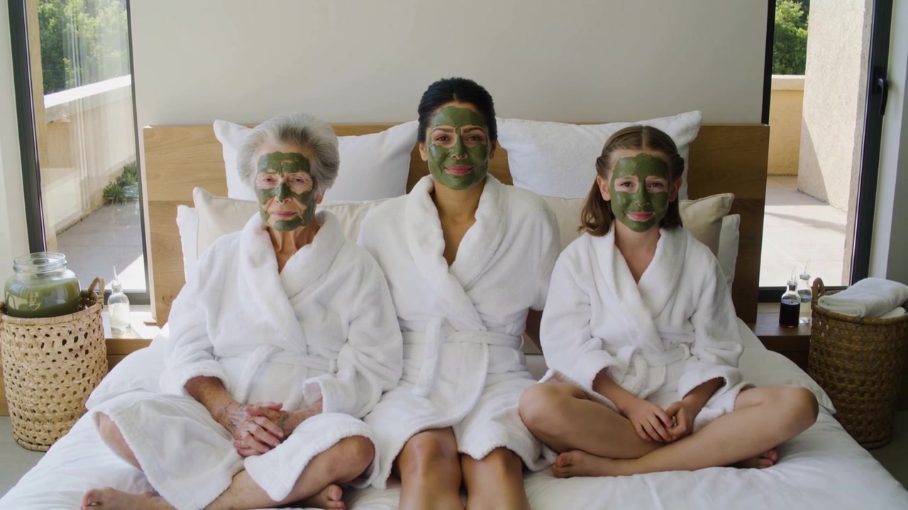 Tre kvindelige familiemedlemmer iført hvide badekåber deler spa-tid i soveværelset med ansigtsmaske.