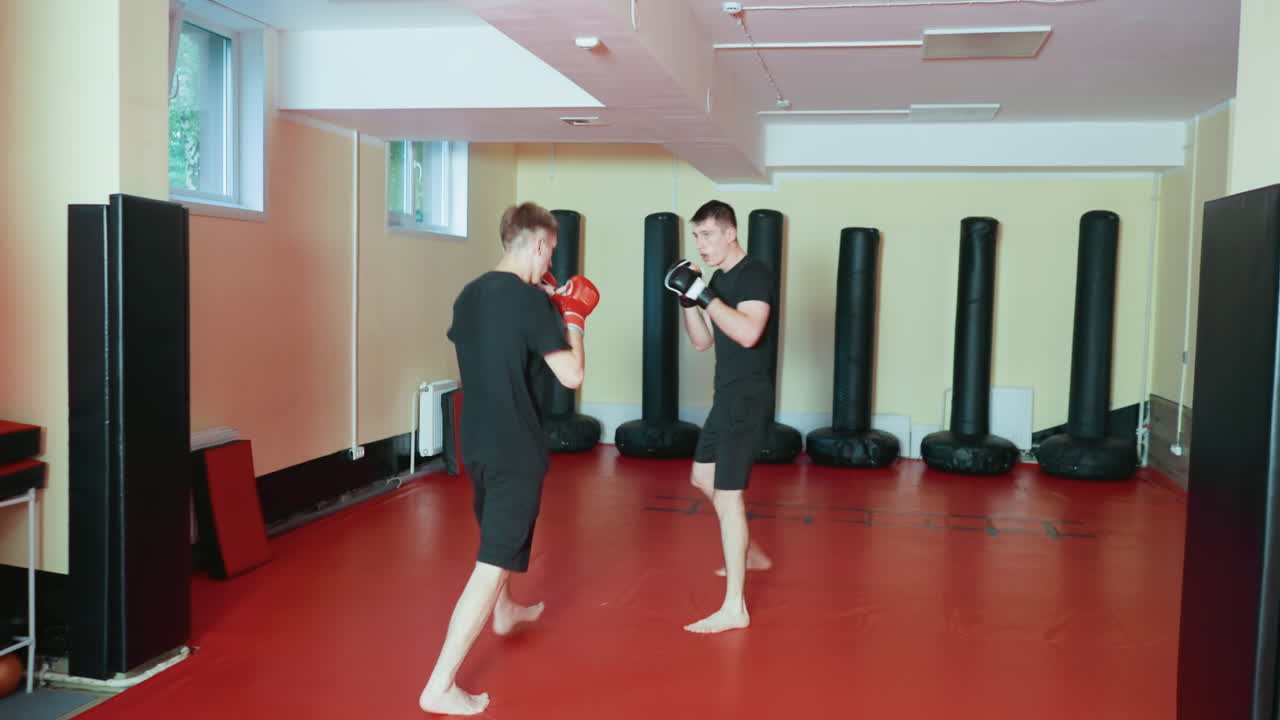 Two wrestlers face off in sparring session inside training gym, barefoot on red mat, wearing black sportswear and gloves, preparing for combat practice surrounded by tall punching bags against yellow walls