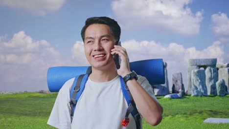 close up of asian male hiker with mountaineering backpack smiling and talking on smartphone while traveling in stonehenge