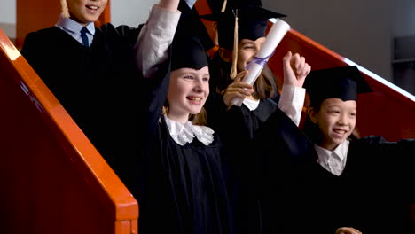 Gruppe-Glücklicher-Vorschulkinder-In-Mütze-Und-Mantel,-Die-Auf-Treppen-Stehen-Und-Ihre-Abschlusszeugnisse-Zeigen