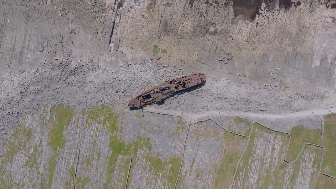La nave da carico Plassey arrugginita giace arenata su una costa rocciosa spoglia mentre la telecamera si libra direttamente sopra in pieno giorno.