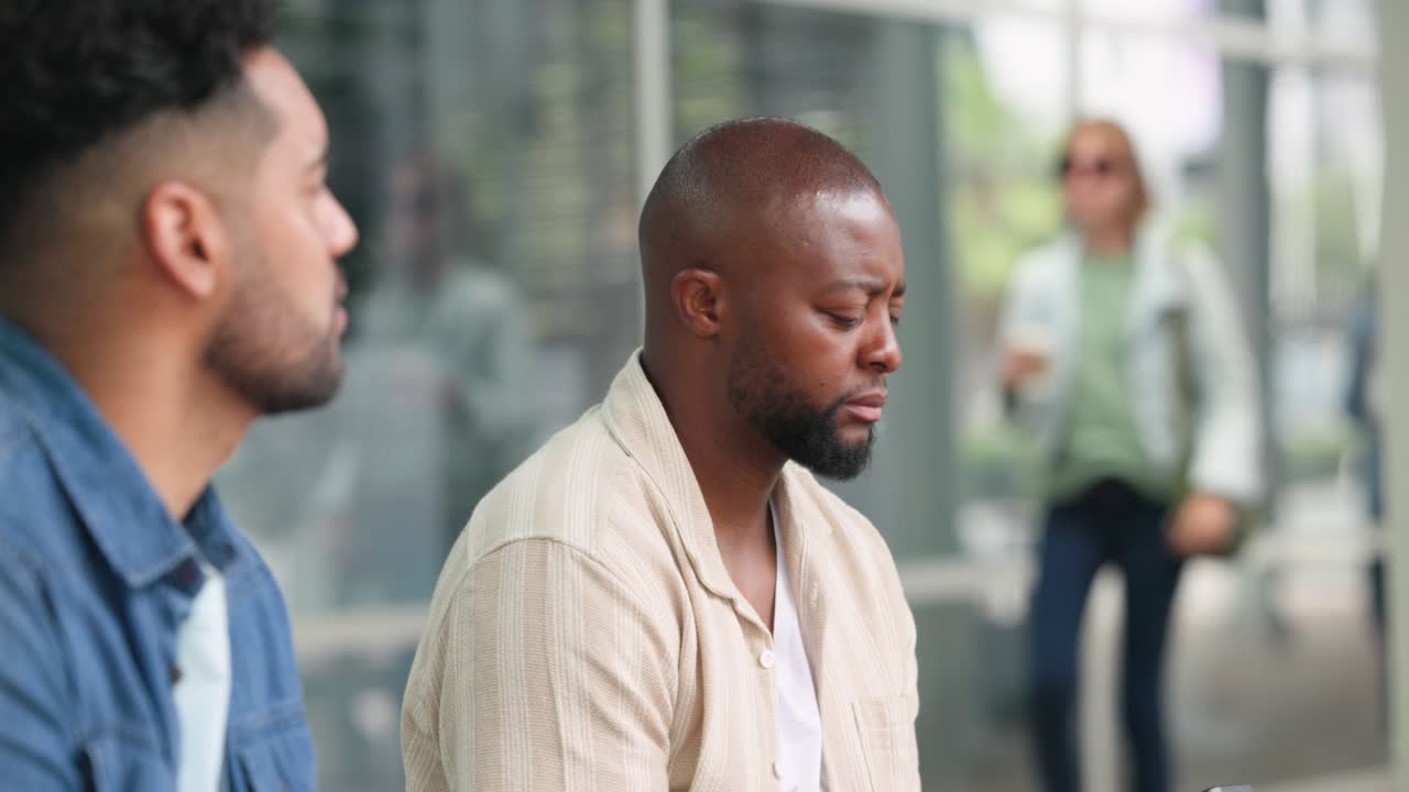 Due uomini su una panchina in piazza, un uomo afroamericano che esamina e usa il telefono mentre aspetta, con la folla che passa.