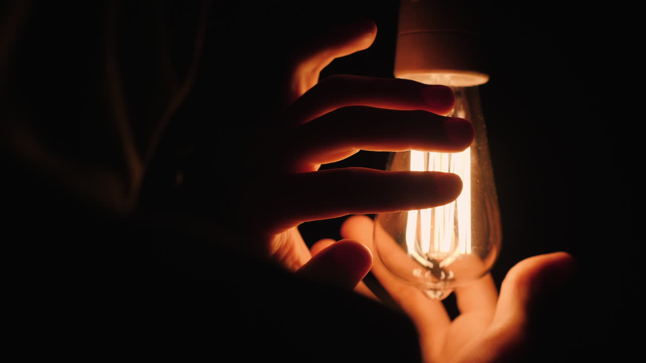 Back View Of Man Warms His Hands Near A Light Bulb In The Dark