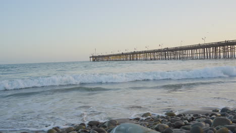 Toma-Panorámica-De-Las-Olas-Al-Atardecer-Rodando-A-Través-Del-Océano-Pacífico-Con-El-Muelle-De-Ventura-En-El-Fondo-Ubicado-En-El-Sur-De-California