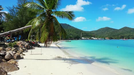 Palm-tree-with-green-leaves-bent-over-white-sand-of-exotic-beach-washed-by-turquoise-lagoon-under-bright-blue-sky-with-white-clouds-in-Thailand