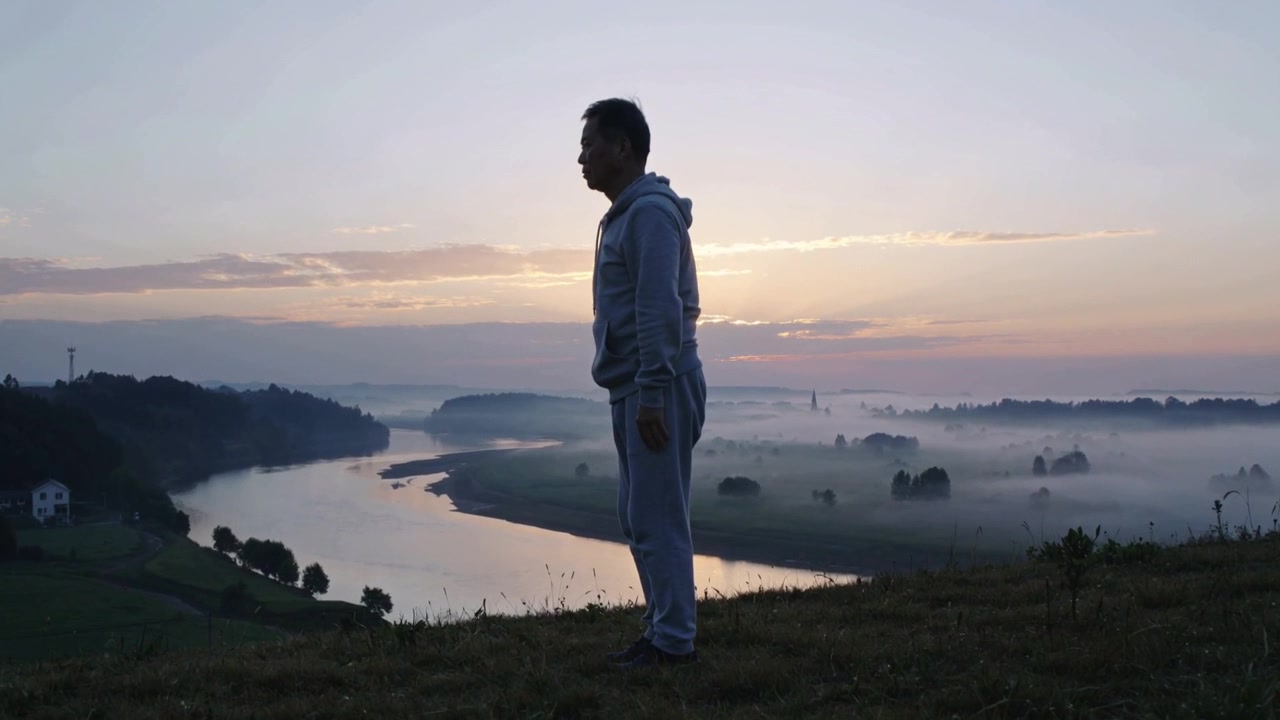 Uomo adulto che si ferma dopo aver fatto jogging, si allunga su una collina erbosa all'alba, con nebbia sul fiume e felpa con cappuccio.
