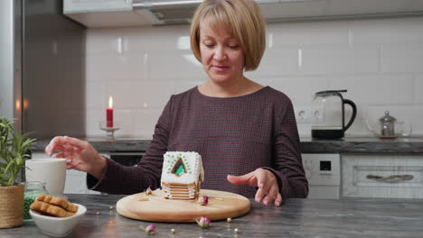 woman in checkered gown sets down mug while carefully rotating decorated gingerbread house on wooden tray, cozy kitchen setting with candlelight, cookies and green beads