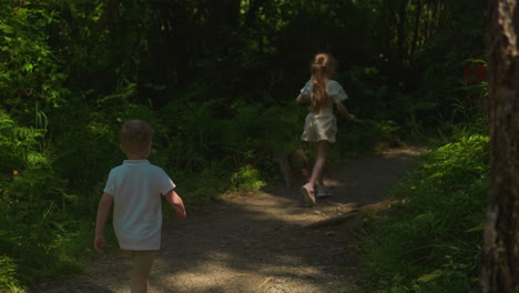 couple of children moves along shaded forest path backside view. preschooler boy and schoolgirl rest actively on vacation slow motion. leisure in nature