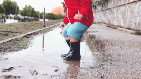 kid wearing rubber boots jumping in a muddy puddle and splashing water in slow motion