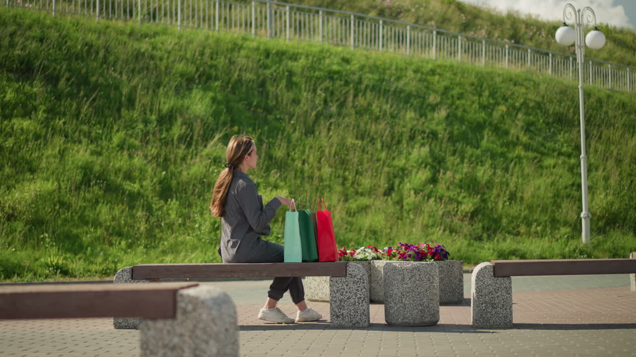 여자는 벤치에 걸어, 다채로운 쇼핑 가방을 떨어뜨리고, 좌석에 앉아, 왼손을 편안하게 하고, 오른쪽 다리를 앞으로 움직여서 멀리 바라보고, 언덕의 초록색이 배경에 보인다.