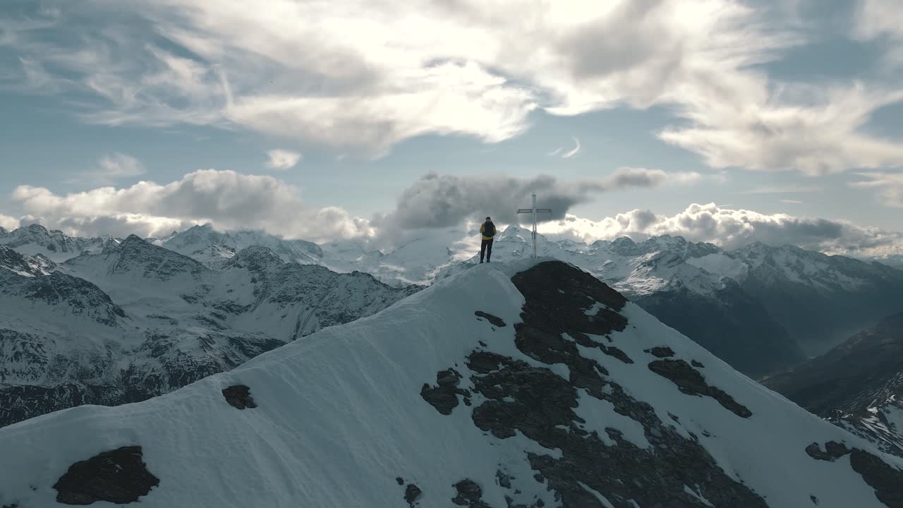 A person stands next to a cross on a summit and enjoys the panorama