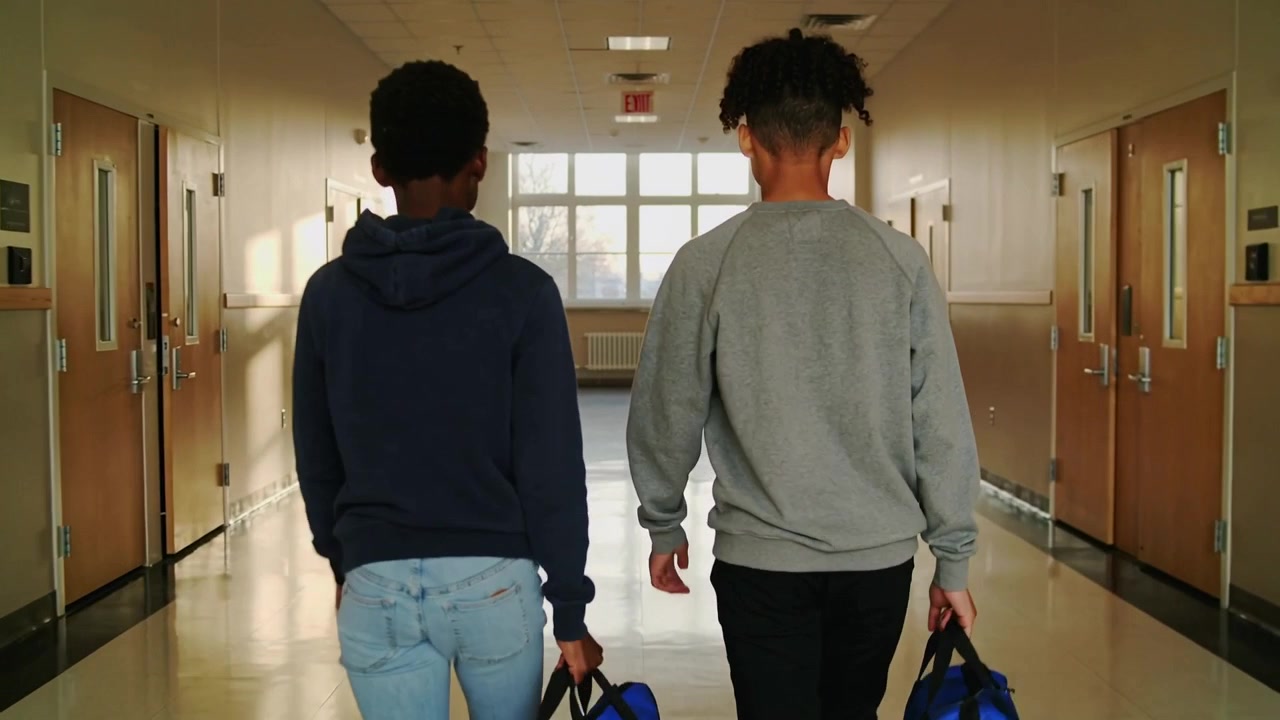 Due adolescenti che camminano, con felpa con cappuccio e felpa, con borsoni blu-neri, verso la finestra del corridoio della scuola.