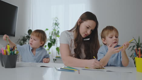 A-young-mother-with-two-children-talking-on-a-mobile-phone-draws-with-a-pencil-and-helps-children-draw-with-colored-pencils