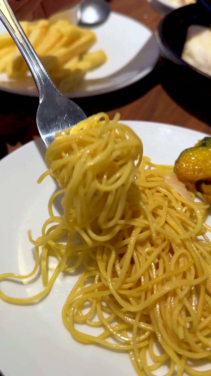 Primo piano ripreso a mano di una forchetta che avvolge spaghetti su un piatto bianco sotto la calda illuminazione di un ristorante